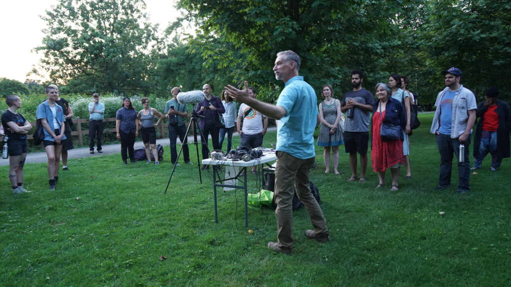 An image of Eric Leonardson welcoming a group of people outdoors to listen