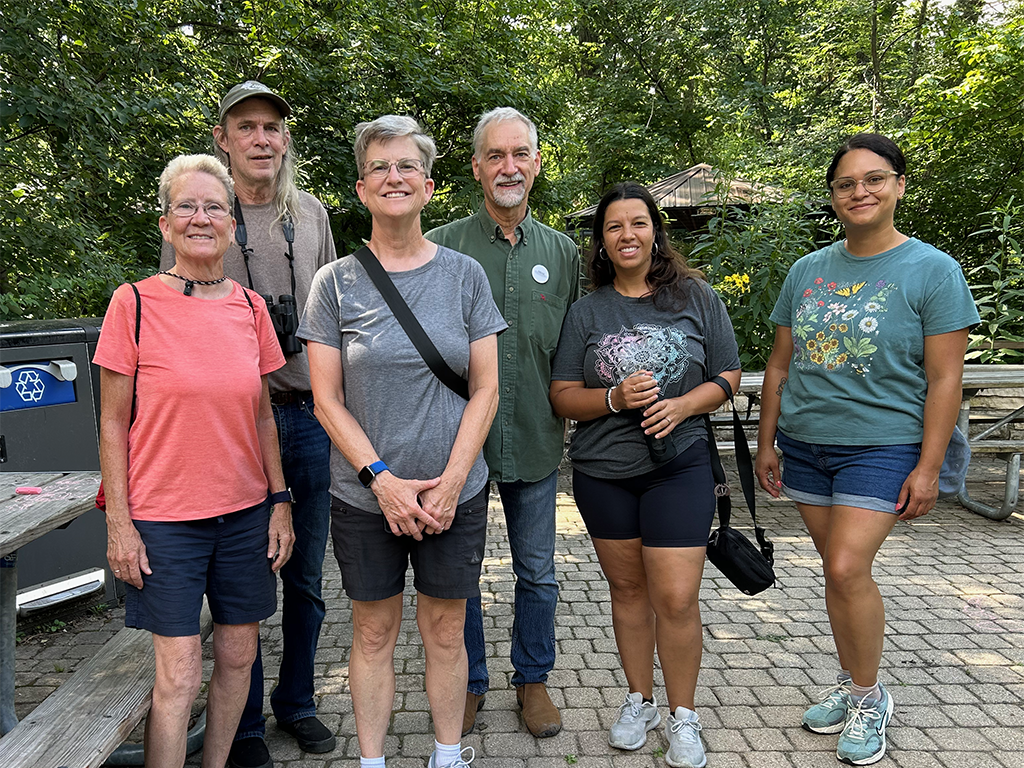 Group of six soundwalk participants smiling together outdoors at a nature center, with trees, picnic tables, and a gazebo roof in the background. One participant carries binoculars, suggesting a nature-listening outing.