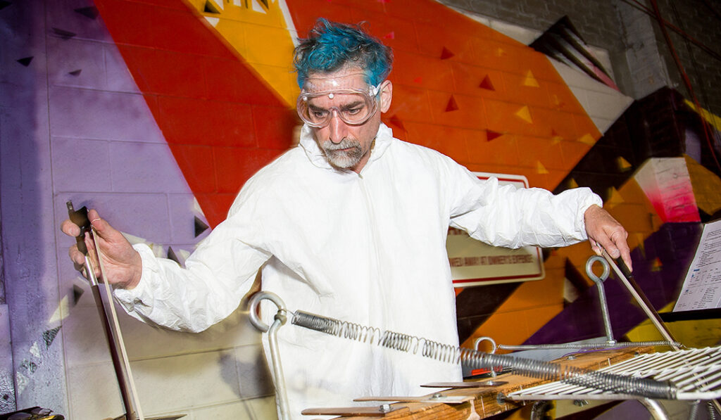 A performer with bright blue hair, safety goggles, and a white protective coverall plays an experimental percussion instrument made of metal rods, springs, and wooden pieces against a brightly painted geometric wall.